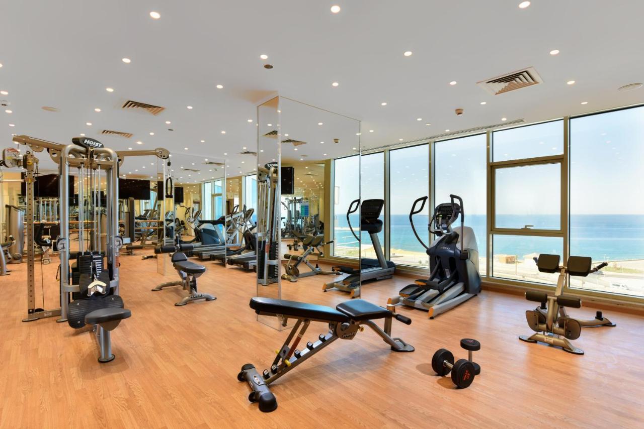 Tolip Resort El Galala Majestic Αΐν Σούχνα Εξωτερικό φωτογραφία The photo shows a modern gym interior with a variety of fitness equipment. There are cardio machines like treadmills and ellipticals, along with strength training equipment such as free weights and benches. The gym features large windows that offer a
